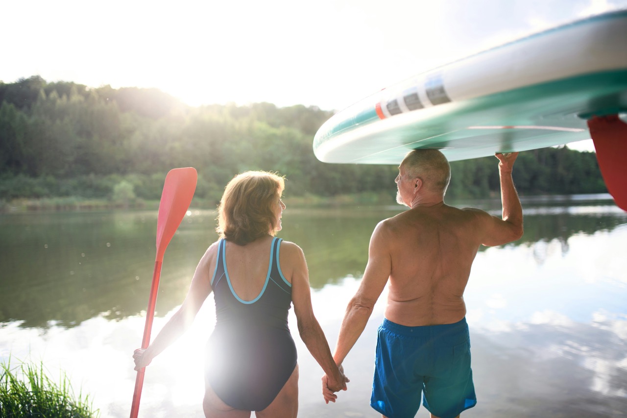 senior couple carrying paddleboard by lake in summer